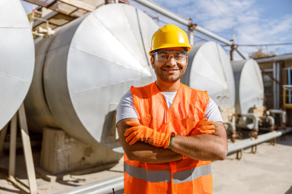 Happy worker in safety equipment, standing in front of storage tanks with crossed arms at heavy industry plant